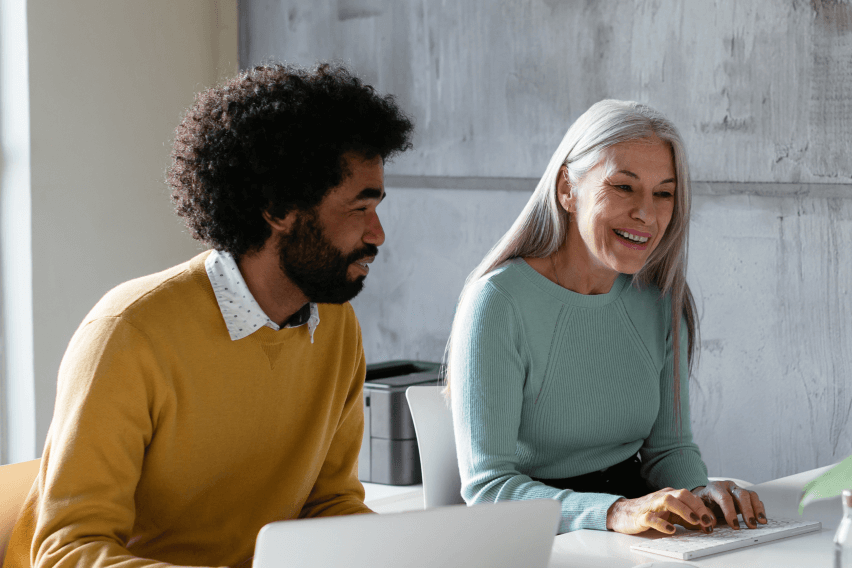 Man and woman sitting together at desk looking at screen while smiling