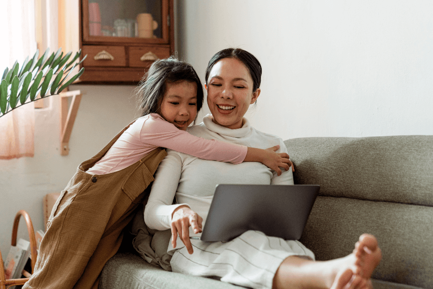 Woman works on laptop while her young daughter hugs her
