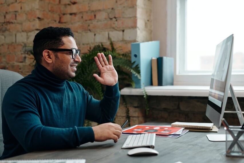 Man at desk on video call, raising hand in greeting