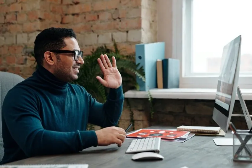 Man at desk on video call, raising hand in greeting