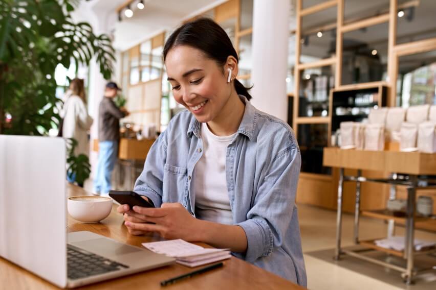 smiling woman with laptop working in coffee shop looking at cell phone